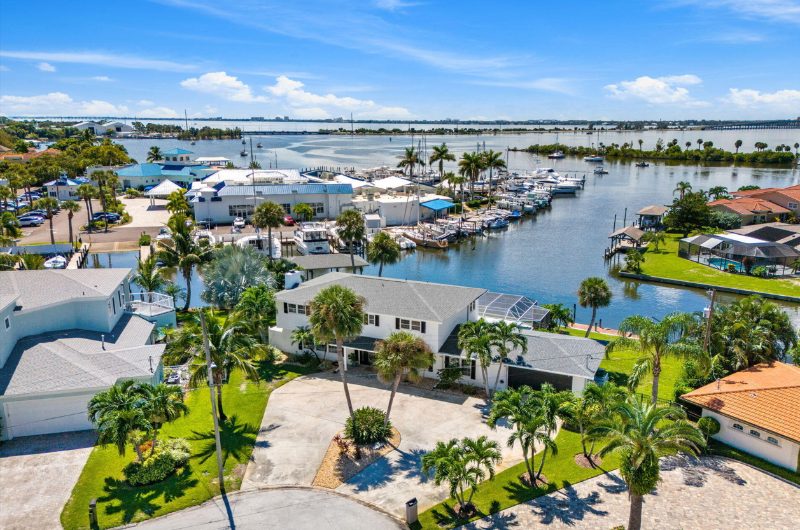 Aerial view of a waterfront home with palm trees, a circular driveway, and boats docked in the nearby harbor.